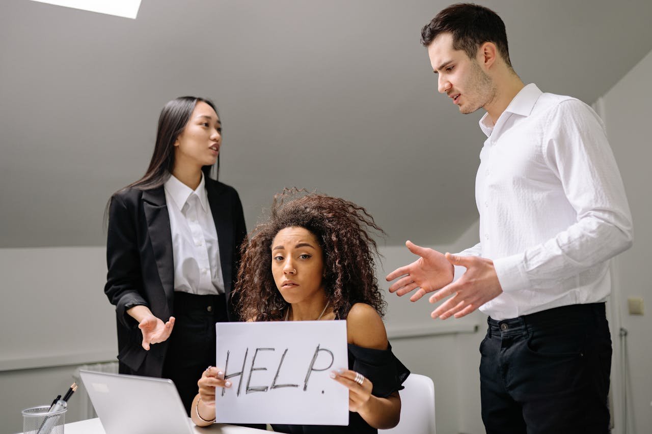 A woman in an office holds a HELP sign while facing conflict with coworkers.